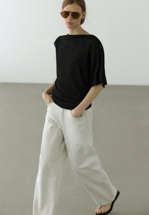 Kvinde iført oversized sort top, brede hvide bukser, brune solbriller, sorte flip-flops, gående med hænderne i lommerne på gråt gulv.