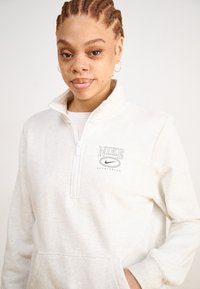 Lys grå Nike zip-up hettegenser med høy krage, frontlomme og brodert logo. Myk tekstur med avslappet passform.