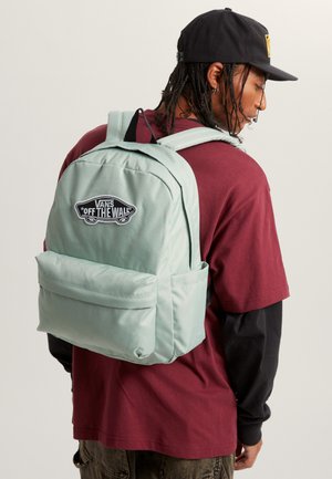 Zaino Vans verde chiaro con tasca frontale, spallacci imbottiti e patch con logo. Il tessuto appare liscio e resistente. Indossato con una maglia bordeaux.