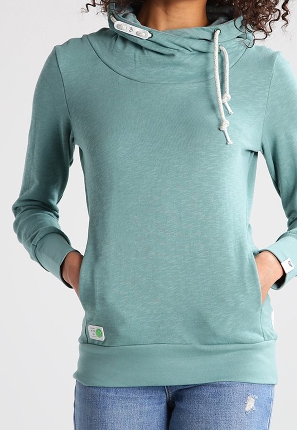 Femme portant un sweat à capuche bleu sarcelle avec cordons blancs et poches, associé à un jean bleu déchiré, debout les mains dans les poches.