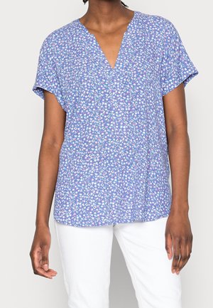 Vrouw die een blauwe korte mouwen bloes met bloemenprint en V-hals draagt, gecombineerd met een witte broek, staand voor een effen achtergrond.