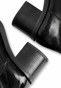 Mustat kiiltävät nahkaiset nilkkurit leveillä pinoiduilla koroilla, lähikuvassa valkoisella taustalla.