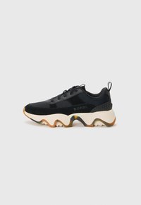 Sorte treningsko med teksturert mesh og suede-materialer, med en chunky hvit såle med zigzag-design og en trekkfane på hælen.