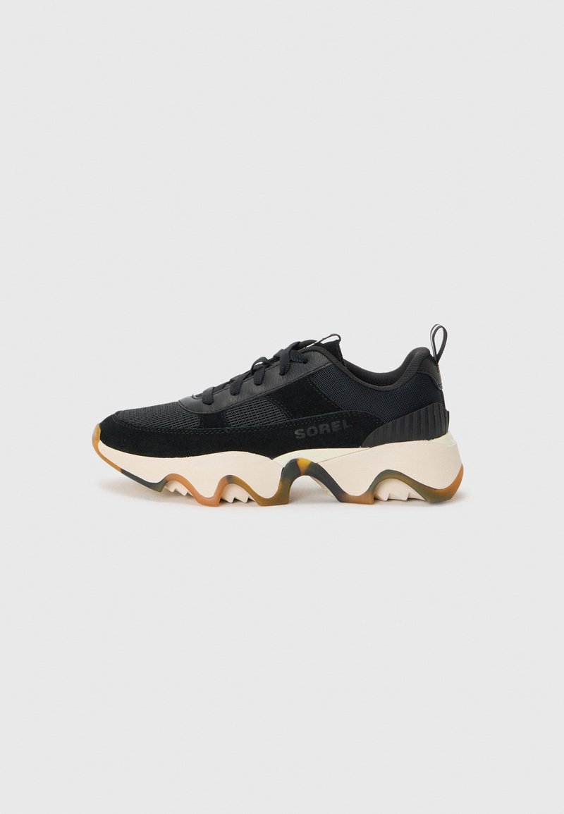 Sorte treningsko med teksturert mesh og suede-materialer, med en chunky hvit såle med zigzag-design og en trekkfane på hælen.