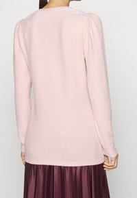 Ljust rosa pullovertröja med långa ärmar, mjuk textur och spetsdetalj vid axeln, tillsammans med en mörk veckad kjol.