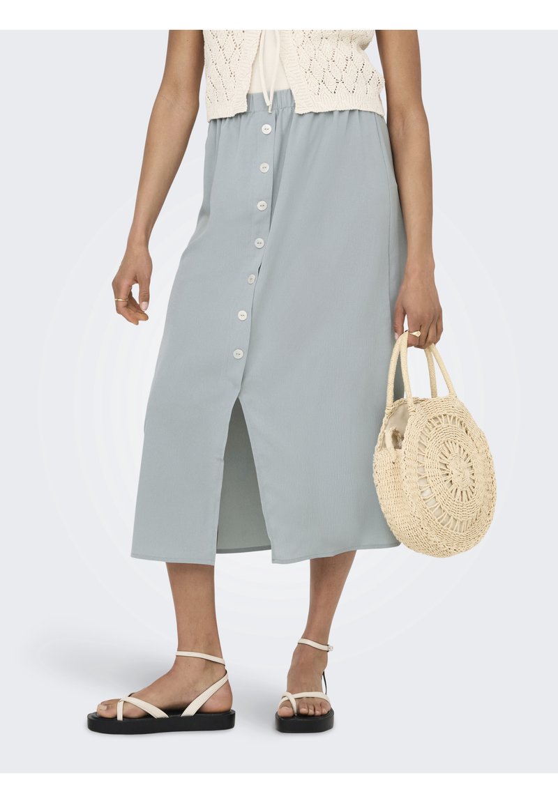 Femme portant une jupe bleu clair à boutons sur le devant, un haut en maille blanc, des sandales blanches à brides, et tenant un sac à main rond tissé beige.