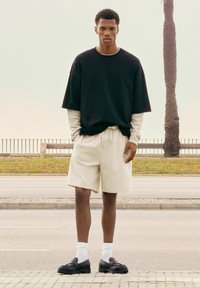 Mlad moški stoji na pločniku, oblečen v črno preveliko majico čez dolg rokav, bež kratke hlače, bele nogavice in črne loaferje, zraven je palmova drevo.