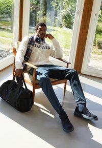 Marineblaue Wildleder-Stiefeletten, marineblaue Hose, cremefarbener Pulli mit Zopfstrick und Logo, gestreifter Schal, schwarze Reisetasche, sitzend auf einem Holzstuhl.