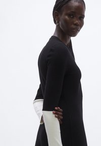 Čierne šaty s dlhými rukávmi a priliehavým strihom, s kontrastnými bielymi manžetami. Textúra pôsobí hladko a dizajn je minimalistický.