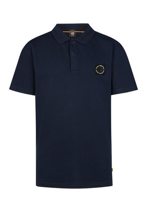 Tienerjongen staand, dragend een marineblauw poloshirt, blauwe spijkerbroek en witte sneakers tegen een effen lichte achtergrond.