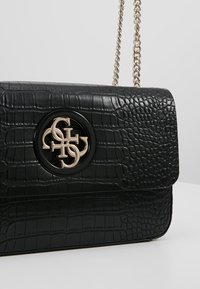Černá kožená kabelka se vzorem krokodýlí kůže, zlatým kruhovým logem a řetízkovým popruhem. Má překlápěcí zapínání a hladký povrch.