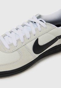 Bijela platnena i nubuk tenisica s crnim Nike logom, bijelim vezicama i crnim gumeni potplat. Sadrži glatke i teksturirane materijale.