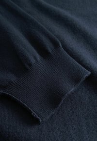 Poignet de pull en maille bleu marine foncé avec texture côtelée, posé sur un tissu assorti.