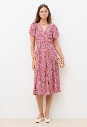 Femme portant une robe midi rose à fleurs avec des manches bouffantes, tenant une pochette beige, chaussée de talons bas blancs et portant des bijoux en or, debout devant un mur blanc.