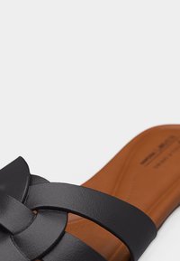 Κοντινό πλάνο μαύρου δερμάτινου σανδαλιού με πλεκτά πάνω λουριά και καφέ μαλακό πέλμα.