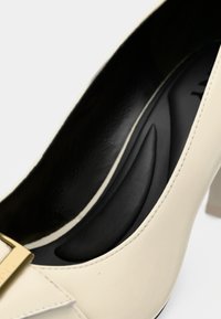 Beige high-heeled shoe with a glossy finish, featuring a black, padded interior and a gold buckle accent. Square heel design.