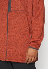 Giacca rossa con zip, dalla texture particolare, caratterizzata da un accento grigio sul polsino e una tasca laterale. Il materiale sembra morbido e dallo stile informale.