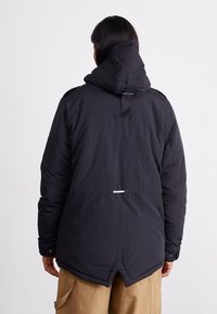 Schwarze isolierte Jacke mit Kapuze, mittellang, mit geknöpften Ärmeln und verstellbarem Taillenzug. Strukturierter Stoff.