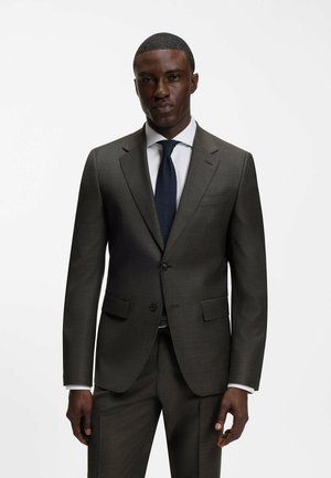 Man staat frontaal gekleed in een donkergrijs pak met bijpassende broek, wit overhemd en een donkerblauwe stropdas tegen een effen witte achtergrond.