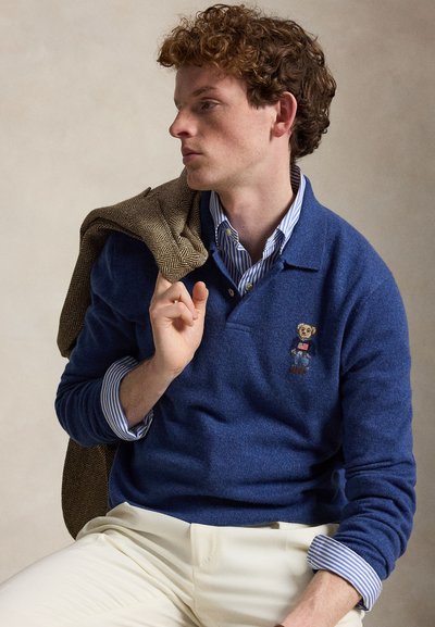 Junger Mann mit lockigem Haar, der ein blaues Langarmhemd mit Bärenemblem trägt und eine braune Fischgrätenjacke über der Schulter hält.