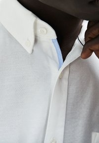 Biała koszula z guzikami, wykonana z teksturowanej tkaniny. Posiada jasnoniebieski akcent pod kołnierzem i kontrastowe czarne przeszycia.
