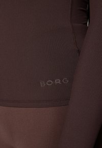 Brun ribbad träningstopp med en slät nederdel, prydd med präglad "BORG"-logotyp. Materialet verkar töjbart och åtsittande.