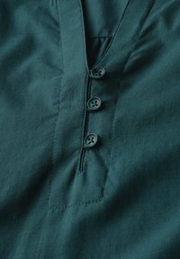 Top in cotone color teal con scollatura a V, caratterizzato da una patta con quattro bottoni scuri e una tasca a toppa sul lato sinistro. Texture liscia.