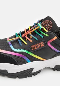 Chaussure de sport noire avec des lacets arc-en-ciel, une surface texturée, des détails en mesh et une semelle blanche. Porte la marque Versace Jeans Couture.