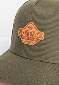 Zaļš cepure ar teksturētu audumu, kurai ir brūns ādas spilventiņš, uz kura ir emblemas formā rakstīts "BLACKSKIES PREMIUM SUPPLIES MMIX".
