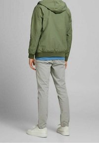 Giacca a vento verde oliva con cappuccio e polsini elasticizzati, indossata sopra una maglietta blu. I pantaloni slim-fit grigio chiaro e le sneaker bianche completano l'outfit.