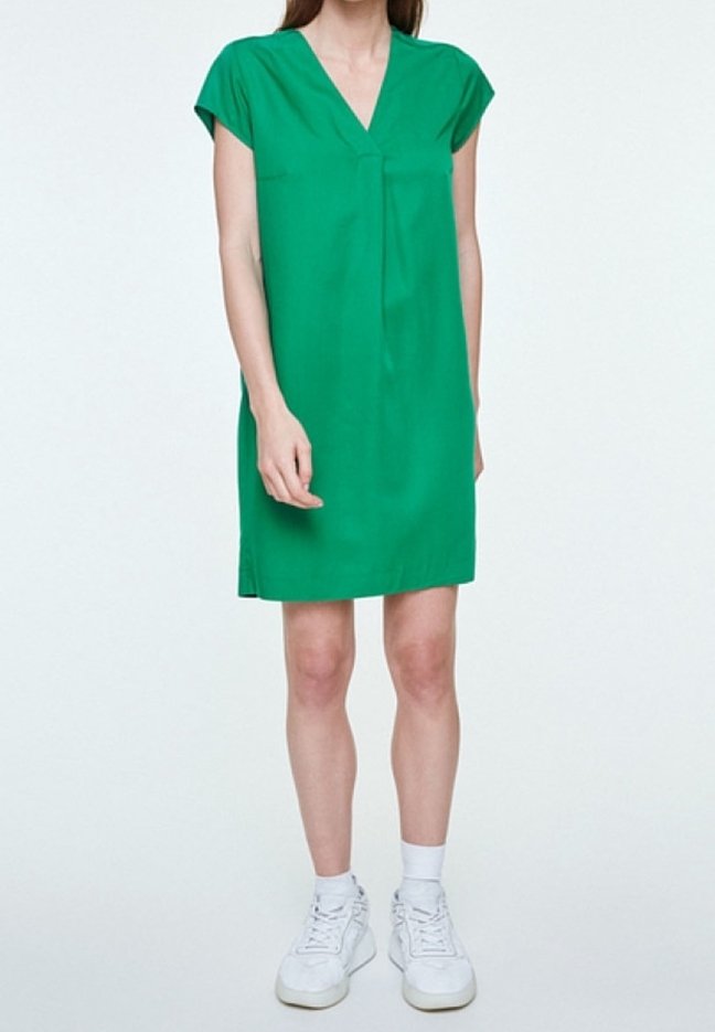 Femme portant une robe verte à manches courtes avec un col en V, des chaussettes blanches et des baskets blanches, debout devant un fond uni.