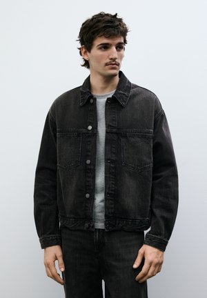 Giovane uomo con capelli scuri e baffi che indossa una giacca di jeans nera e una maglietta grigia, in piedi davanti a uno sfondo semplice di colore grigio chiaro.