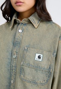 Light blue denim shirt with a pointed collar, silver snaps, and a chest pocket featuring a logo patch. Fabric shows subtle texture and fading.