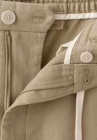 Pantalon beige partiellement déboutonné avec fermeture par bouton, montrant un cordon intérieur blanc et les détails de la texture du tissu.