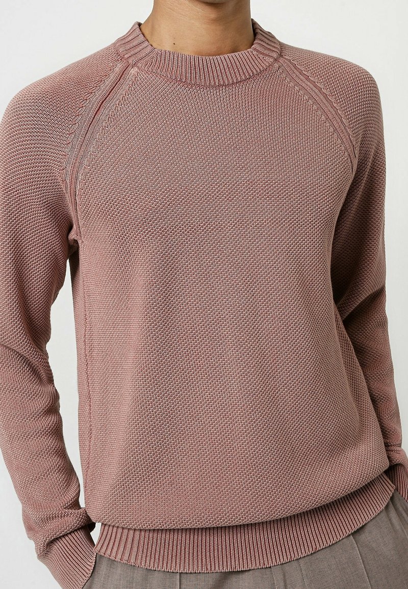 Gros plan d'une personne portant un pull en maille texturée mauve avec col rond, poignets et ourlets côtelés, associé à un pantalon gris.