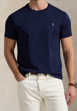 Mann trägt ein marineblaues kurzärmeliges T-Shirt mit kleinem gesticktem Logo, weiße Hose, brauner Gürtel und eine Uhr am linken Handgelenk.