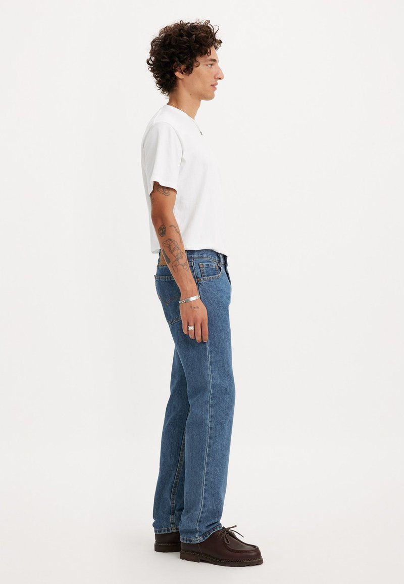 T-shirt en coton blanc, jeans en denim bleu et chaussures marron. La tenue est portée par une personne de profil, mettant en valeur un look décontracté.