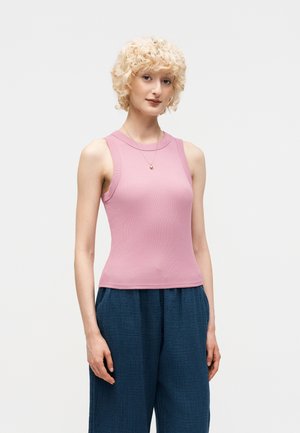 Νέος με σγουρά ξανθά μαλλιά φοράει ρόζ αμάνικη μπλούζα και φαρδιά σκούρα μπλε παντελόνια, στέκεται μπροστά σε απλό ανοιχτό φόντο.