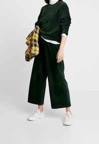 Oversized sweatshirt van groene ribstof en bijpassende brede broek, gecombineerd met witte sneakers. Houdt een gele geruite tas vast.