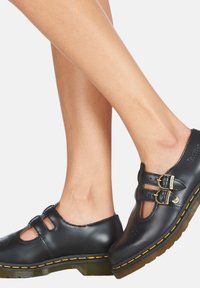 Zapatos de cuero negro con dos hebillas decorativas, detalles recortados y costuras amarillas. Punta redondeada y tacón bajo con suela transparente.