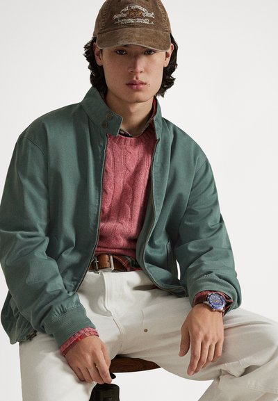 Πράσινο μπουφάν bomber με ριμπ μανσέτες, ροζ πουλόβερ με πλέξη καλώδιο, λευκό παντελόνι και καφέ καπέλο από κορντούρα με λογότυπο. Μπλε ρολόι στον καρπό.