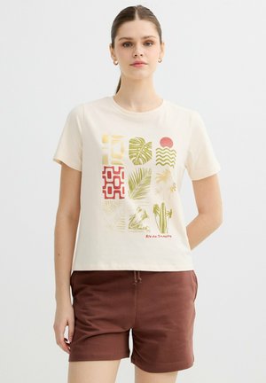 Mujer joven con una camiseta beige con estampados abstractos y tropicales multicolor y pantalones cortos marrones, de pie frente a un fondo blanco liso.
