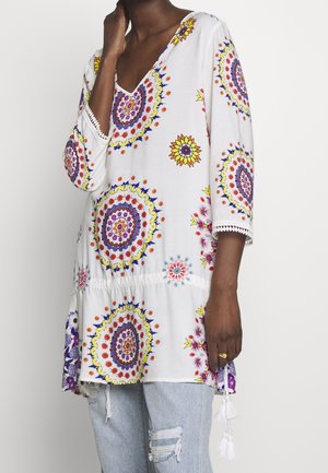 Woman wearing a white tunic with multicolored circular floral patterns and 3/4 sleeves paired with light blue ripped jeans.