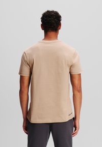 Bežinis t-shirt su apvalia iškirpte ir trumpomis rankovėmis, pagamintas iš lygaus audinio. Yra subtilus logotipas dešiniajame apačioje ir tiesus apatinis kraštas.