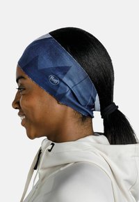 Bandeau bleu marine avec des motifs géométriques, fabriqué en tissu extensible, portant un logo sur le côté et porté avec les cheveux attachés.