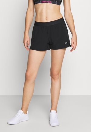 Frau trägt schwarze Calvin Klein Sportshorts und schwarzen Sport-BH, steht in weißen Turnschuhen vor schlichtem, hellem Hintergrund.