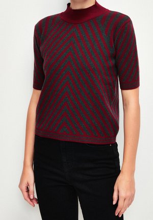 Person iført en kortærmet stribet sweater i mørkerød og mørkegrå med mock-neck, kombineret med sorte bukser mod en ensfarvet baggrund.