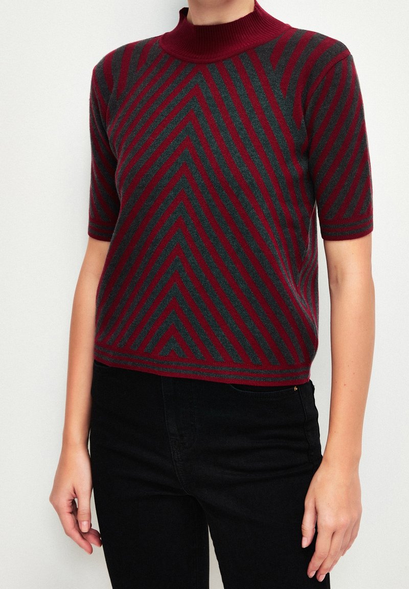 Personne portant un pull à manches courtes rayé bordeaux et gris foncé avec un col montant, assorti à un pantalon noir sur un fond uni.