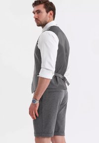 Gilet grigio in tessuto testurizzato abbinato a pantaloni corti grigi. Camicia bianca a maniche lunghe sotto. Cinghia posteriore regolabile, orologio argento al polso.