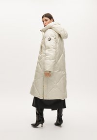 Vadderad lång jacka i ljusbeige med diamantkviltning, en dragsko på huvan samt logotyppatch på ärmen; matchad med svarta knähöga stövlar.